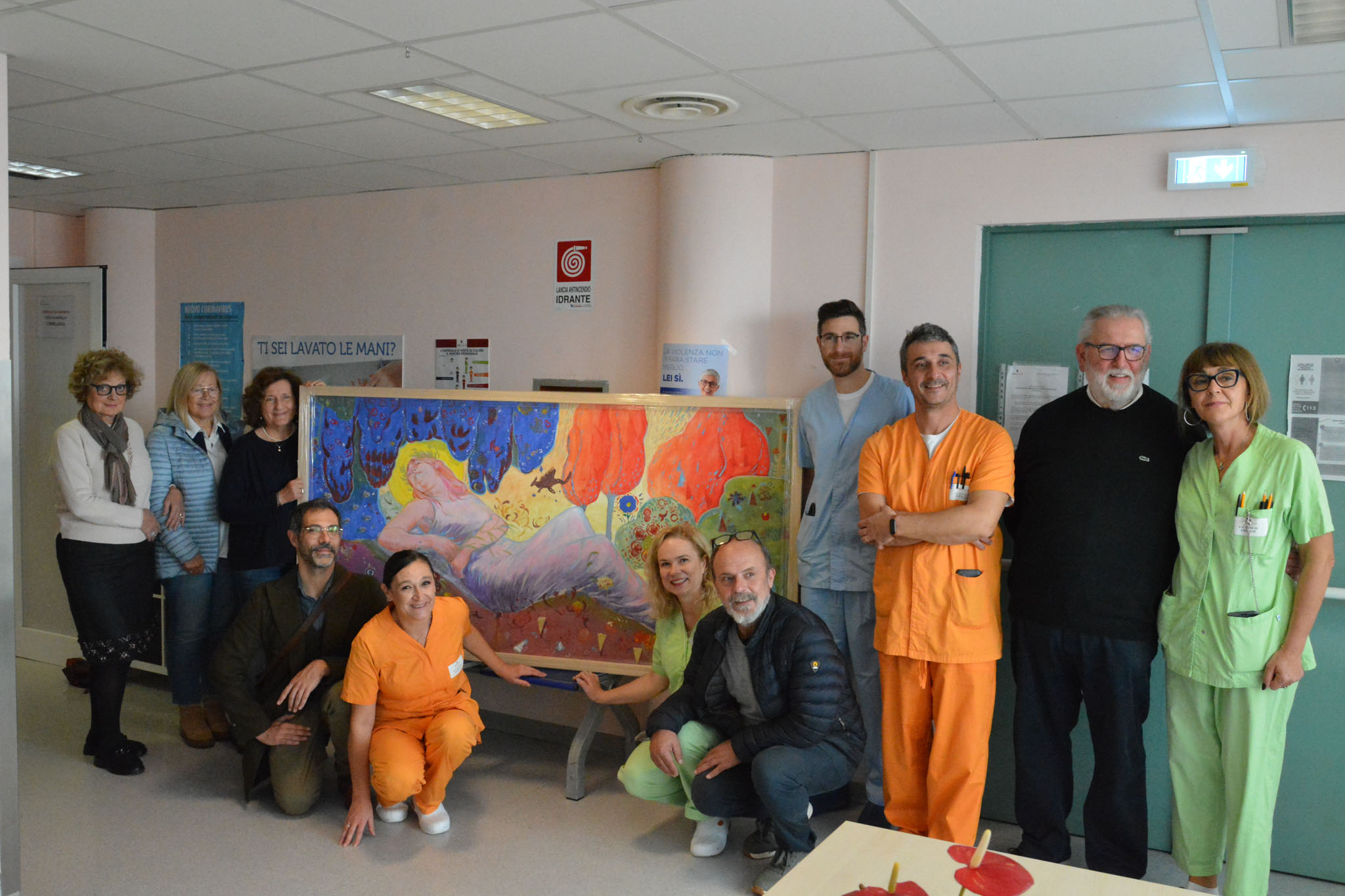 img title Gruppo di persone, tra cui personale sanitario in divise arancioni e verdi e donatori in abiti civili, posano in un corridoio d'ospedale dietro un grande quadro colorato con figure oniriche.