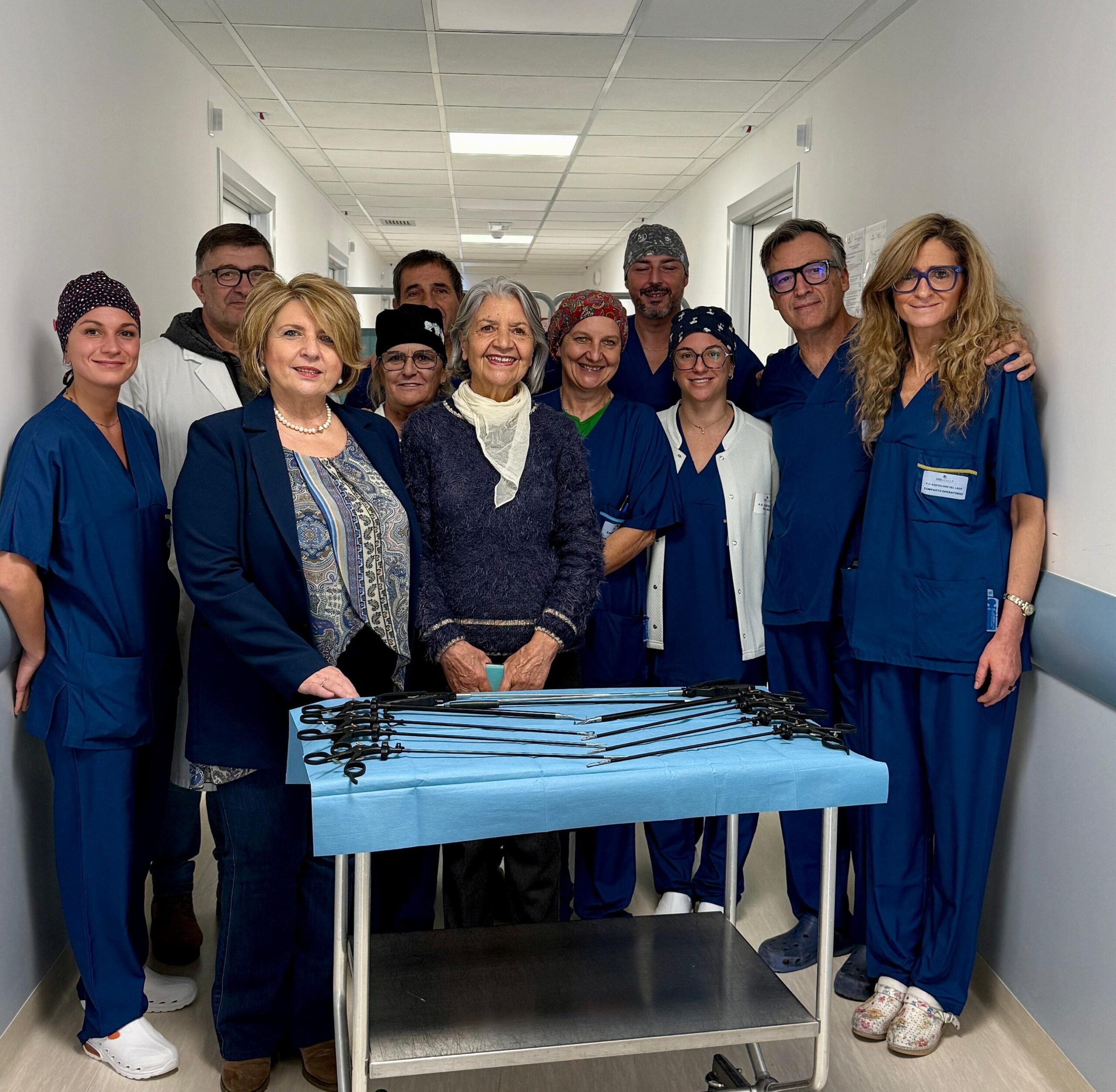 img title Gruppo di medici e infermieri in divisa blu, con due donne in abiti civili, una è la direttrice Teresa Tedesco, posano in un corridoio d'ospedale dietro un carrello con strumenti chirurgici scuri. Celebrazione di una donazione.