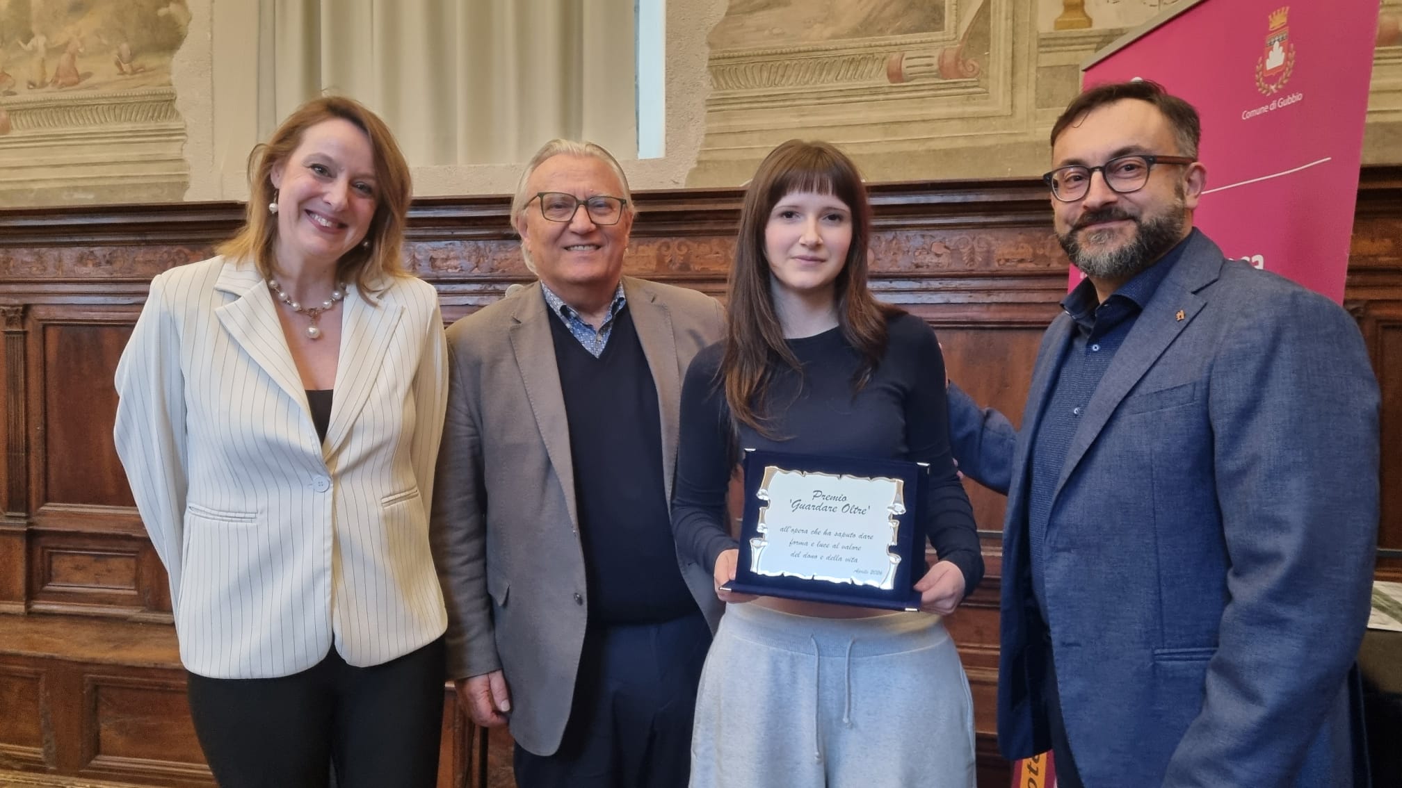 Foto di gruppo durante la premiazione. Quattro persone in posa: a sinistra l'assessore Micaela Parlagreco e il dott. Pierluigi Piergentili; al centro la studentessa vincitrice Jesica Anamaria Rafan che regge una targa blu e argento; a destra il prof. Moreno Panzolini. Sullo sfondo una sala storica con pareti affrescate.
