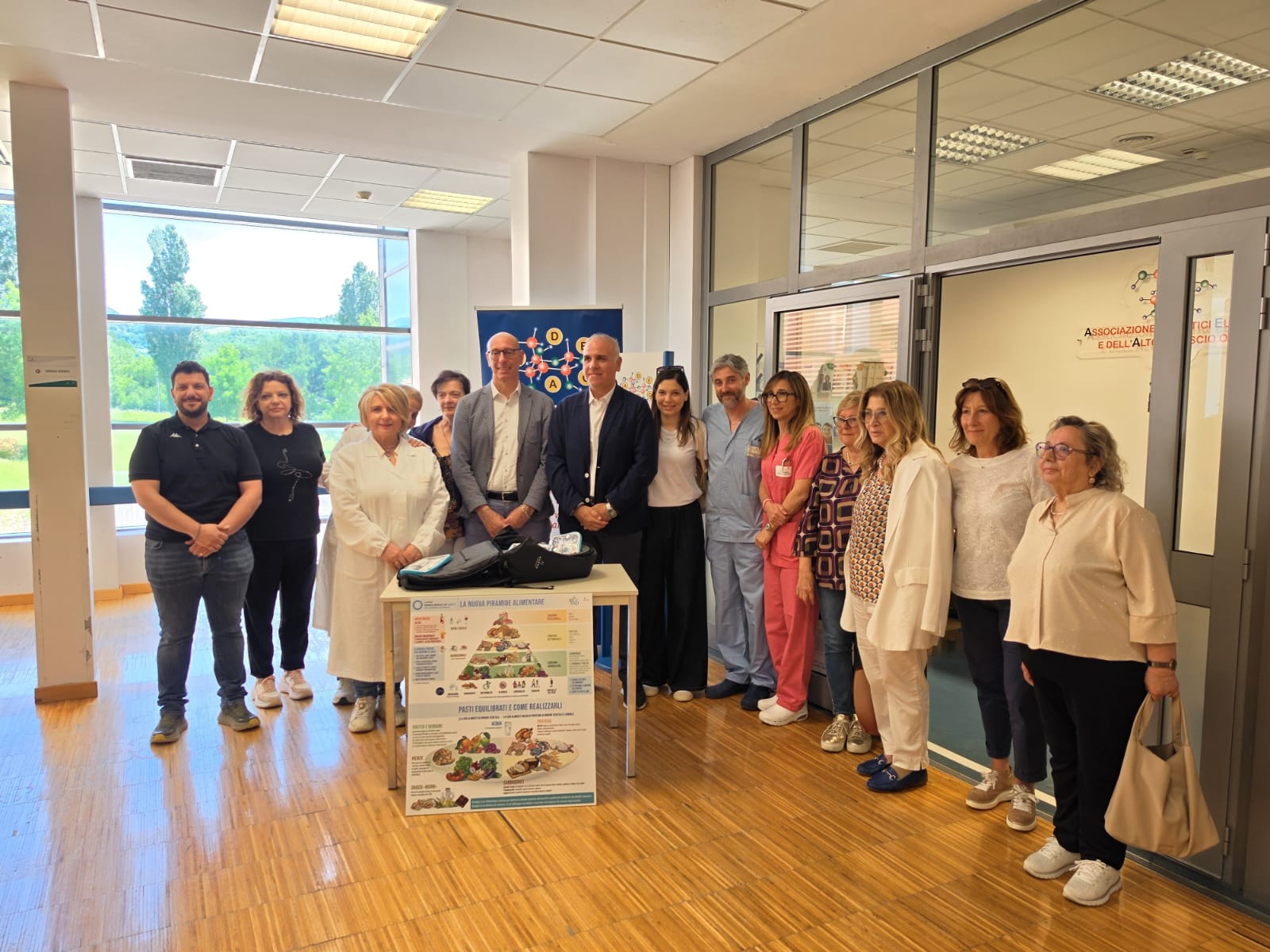 img title Foto di gruppo all'ospedale di Gubbio-Gualdo Tadino per la donazione di un bioimpedenziometro. Autorità e rappresentanti delle associazioni Ade e Correre a Perdifiato posano accanto a un tavolo con lo strumento e un poster sulla piramide alimentare.