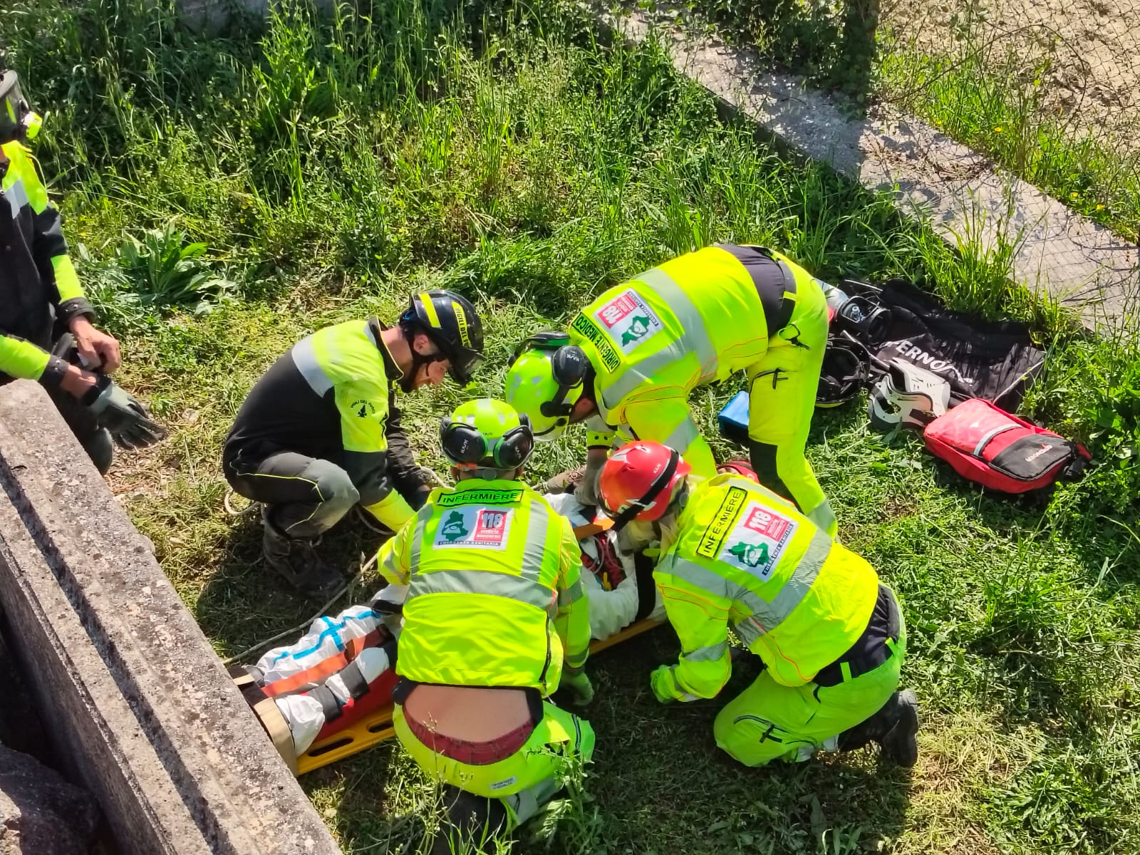 img title Personale sanitario in divisa gialla fluo e Vigili del Fuoco prestano soccorso a un ferito immobilizzato su una tavola spinale gialla in un'area di macerie.