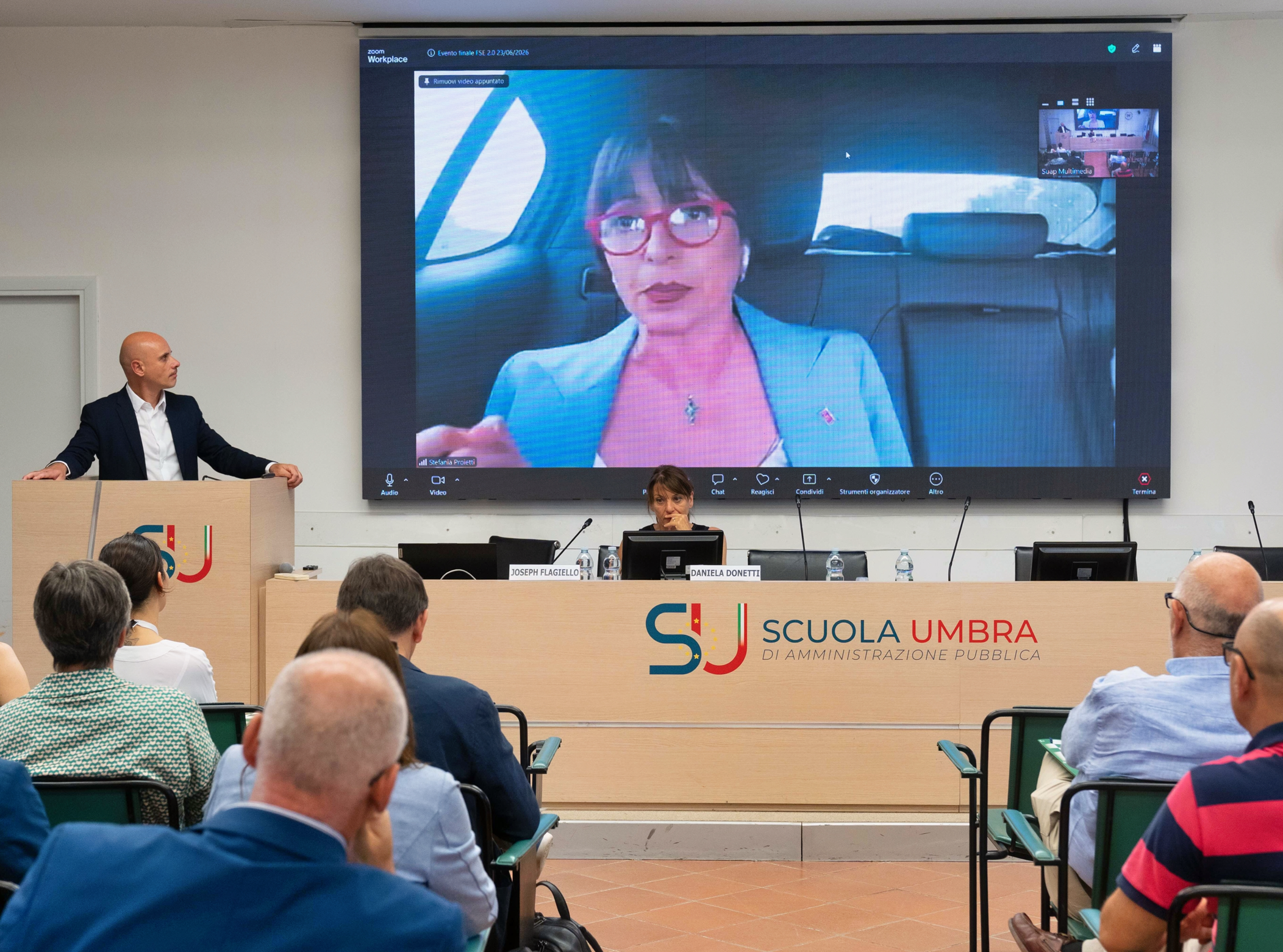 img title Aula magna della Scuola Umbra di Amministrazione Pubblica con un relatore al podio sulla sinistra e la direttrice Daniela Donetti al tavolo della presidenza. Alle loro spalle, un grande maxischermo proietta in videoconferenza Zoom il collegamento in diretta della presidente Stefania Proietti dall'interno di un'autovettura. In primo piano è visibile la platea dei partecipanti seduti.