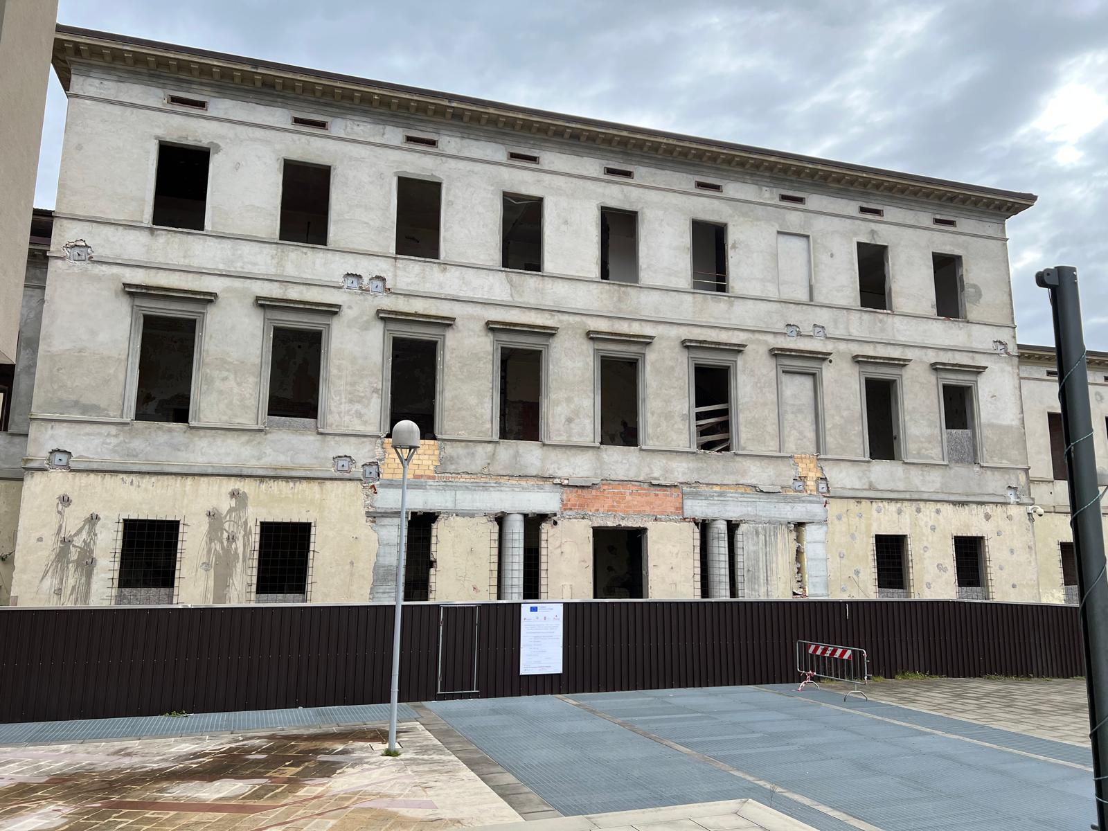 Facciata esterna di un grande edificio storico in ristrutturazione con ponteggi interni e finestre vuote. Davanti alla struttura è presente una recinzione di cantiere scura con un cartello informativo bianco.