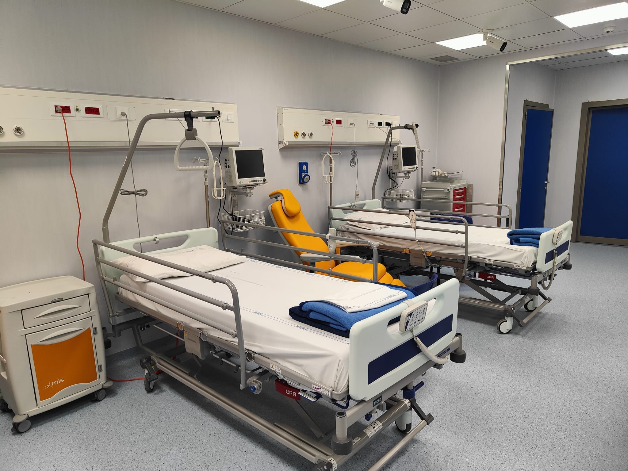 Due letti ospedalieri affiancati in una sala d'attesa attrezzata del Pronto Soccorso, completi di monitor per parametri vitali, poltrona medica gialla e comodino per presidi.