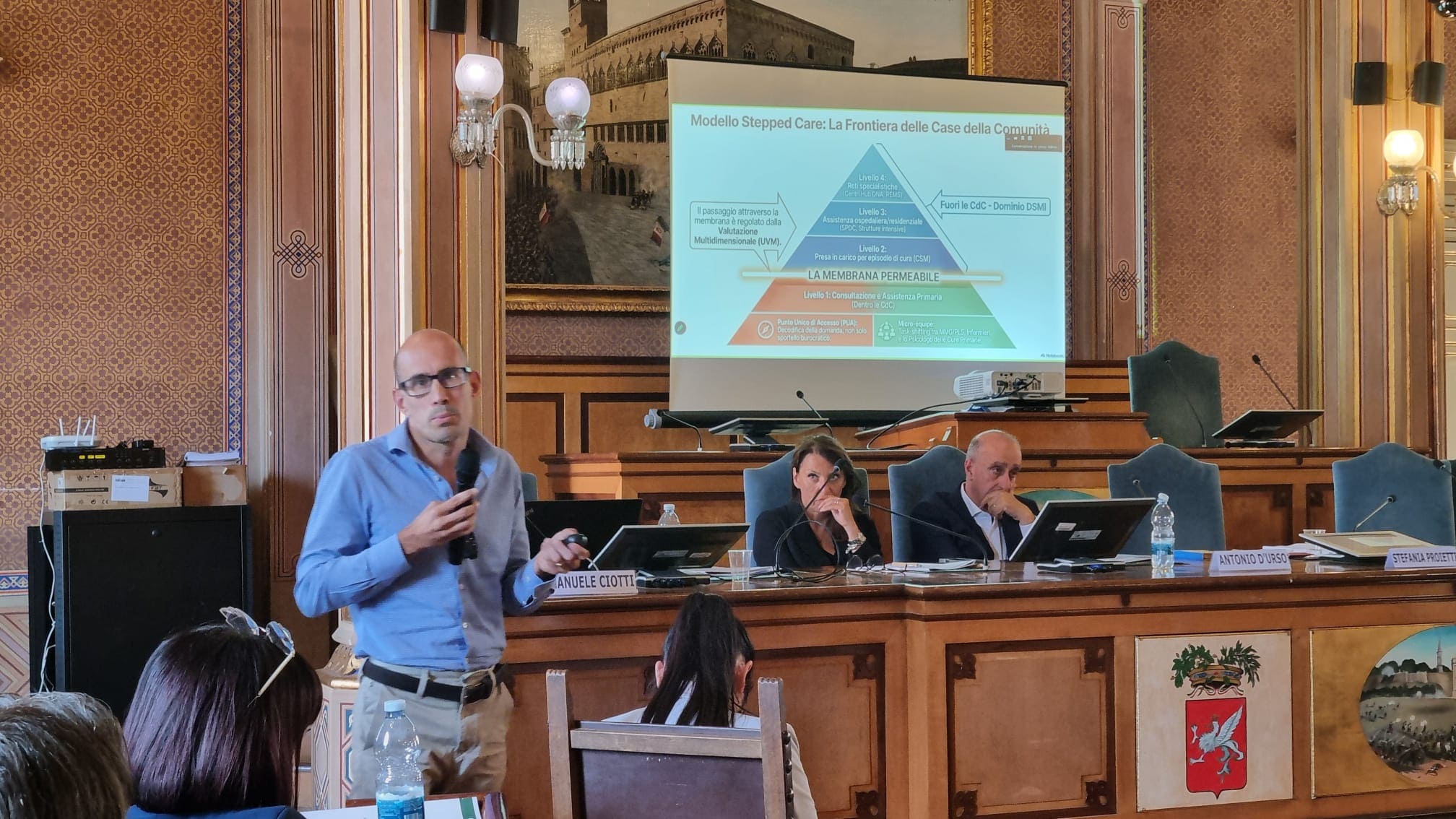 img title Il dottor Emanuele Ciotti in camicia azzurra parla in piedi con un microfono in mano, accanto al tavolo dei relatori; sullo sfondo, lo schermo mostra la piramide del