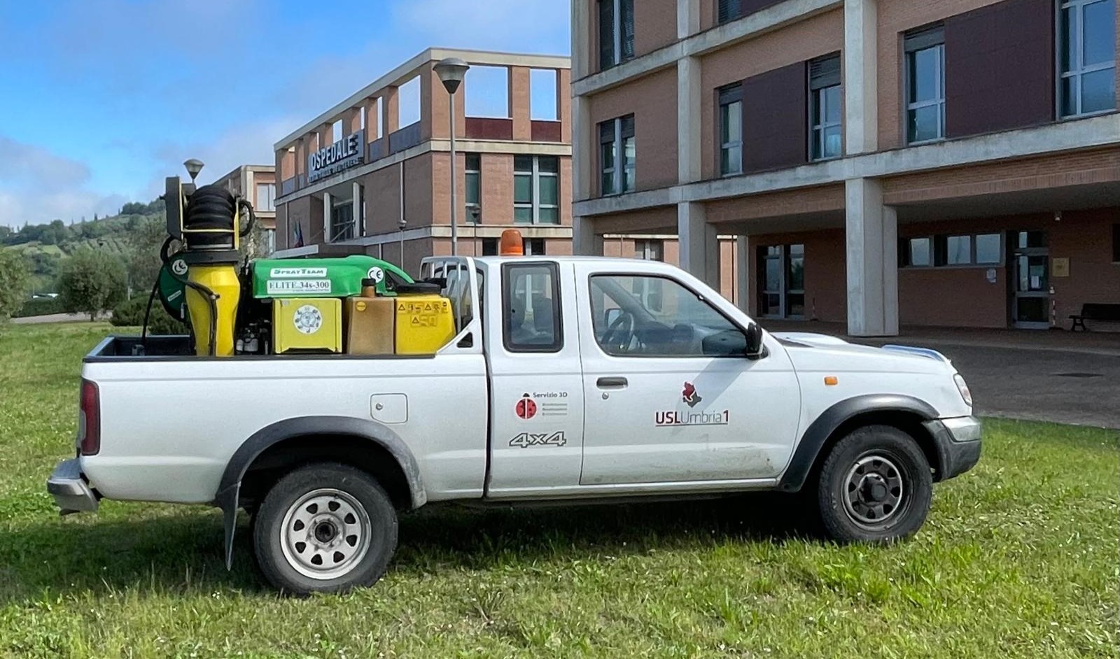 Vista laterale di un automezzo pick-up bianco 4x4 con i loghi ufficiali della USL Umbria 1 e del Servizio sulla portiera, parcheggiato su un prato e allestito nel cassone posteriore con un macchinario attrezzato per la nebulizzazione e la disinfestazione ambientale. Sullo sfondo si vede un edificio ospedaliero in mattoncini.
