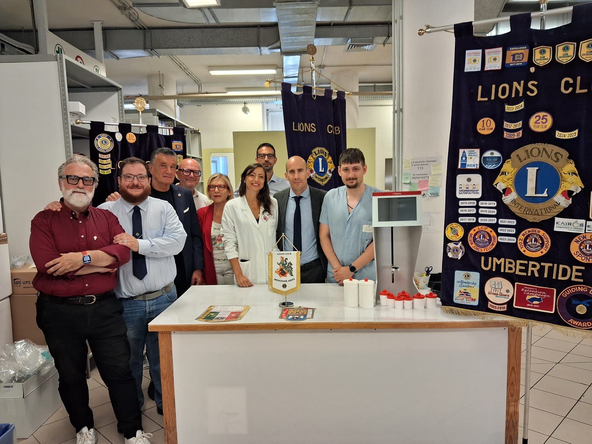 img title Foto di gruppo dei rappresentanti dei Lions Club e della dirigenza sanitaria dell'Usl Umbria 1 riuniti dietro un bancone da laboratorio dove è esposto il nuovo macchinario medico Fagron, affiancato dai gagliardetti e dai labari istituzionali dell'associazione.