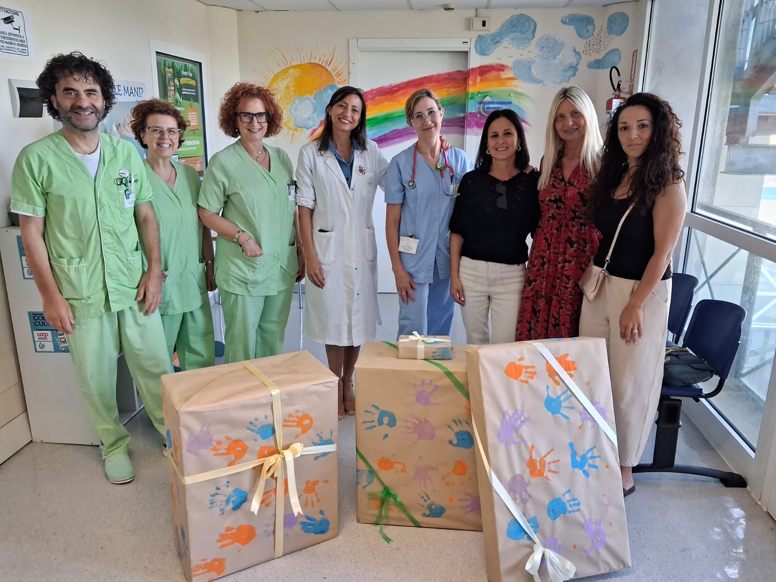 img title Foto di gruppo all'interno del reparto di Pediatria con il personale sanitario in divisa verde e azzurra, la dirigente medica in camice bianco e le rappresentanti della scuola in abiti civili. Davanti a loro, a terra, sono posizionati tre grandi pacchi regalo imballati con carta da pacchi decorata con impronte colorate di mani di bambini e chiusi da nastri. Sullo sfondo si vede un muro dipinto con un arcobaleno e un sole.