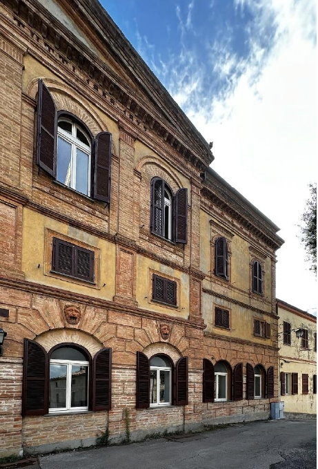img title Vista prospettica dal basso della facciata storica in mattoni dell'ex ospedale di Umbertide, caratterizzata da finestre ad arco con persiane marroni aperte e decorazioni a testa di leone sui portali del piano terra.