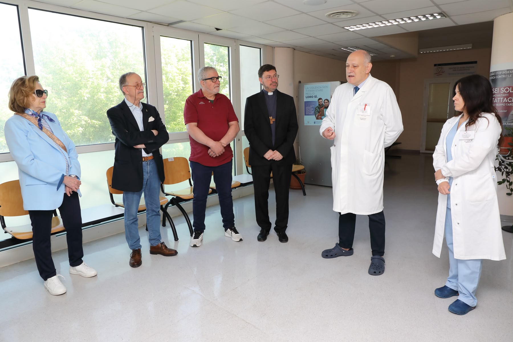 Un gruppo di persone in cerchio nell'atrio del reparto Dialisi ascolta un medico in camice bianco che parla gesticolando. Tra i presenti vi sono esponenti religiosi e civili.