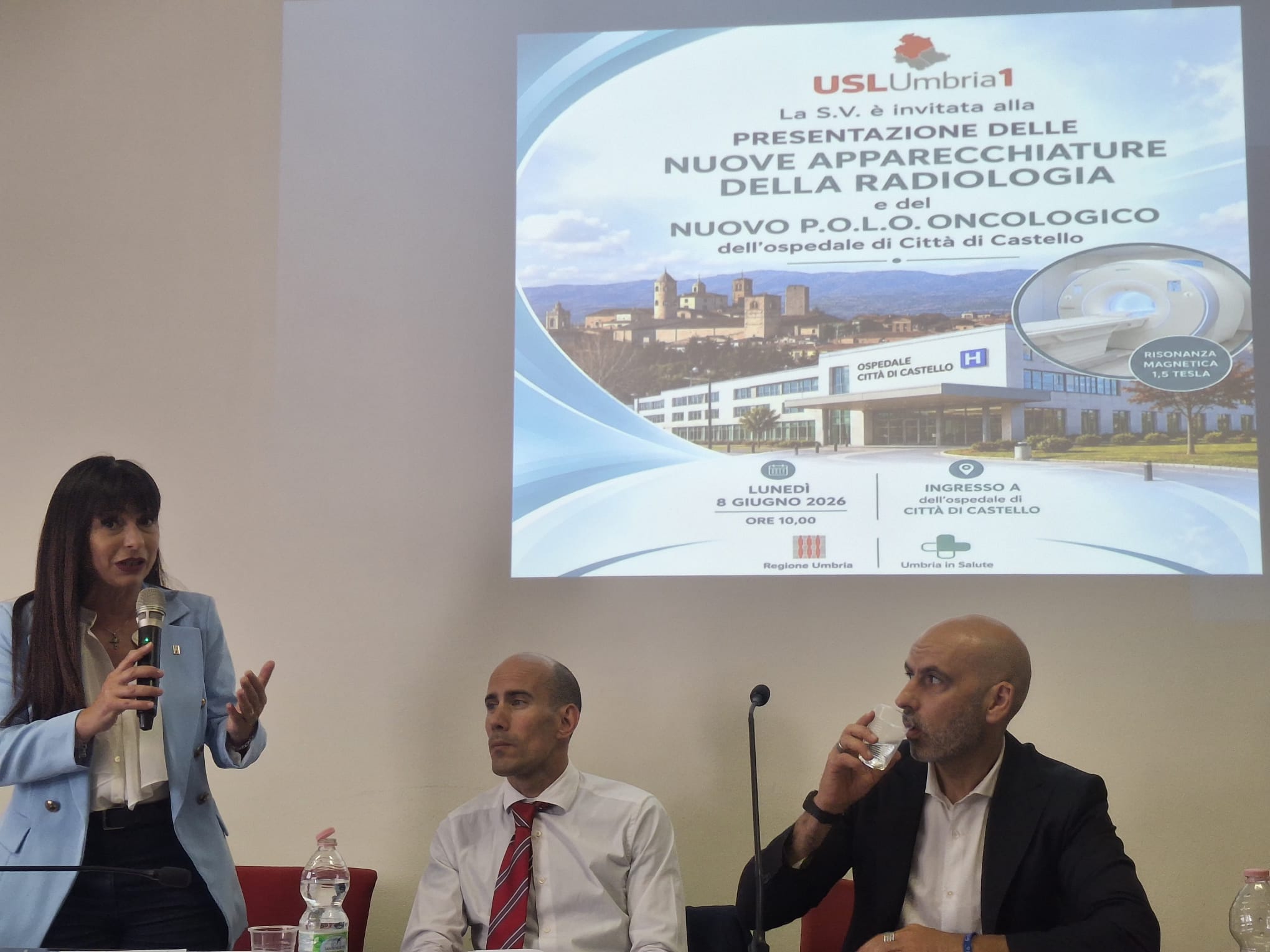 img title La presidente Stefania Proietti parla al microfono accanto al sindaco Luca Secondi e al direttore Emanuele Ciotti, sotto la proiezione dell'invito ufficiale USL Umbria 1 per la presentazione delle nuove apparecchiature di Radiologia e del Polo oncologico del 10 giugno 2026.