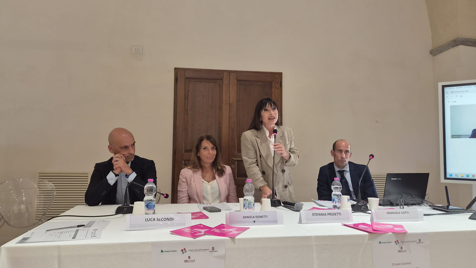 img title Tavolo dei relatori durante un convegno in una sala con pareti chiare e una porta in legno sullo sfondo. Al centro del tavolo, una donna in piedi con giacca beige parla al microfono. Seduti alla sua sinistra ci sono un uomo in abito scuro e una donna con giacca rosa. Alla sua destra siede un altro uomo in abito scuro accanto a un computer portatile e a uno schermo verticale. Sul tavolo sono disposti cartellini con i nomi, bicchieri, bottiglie d'acqua e opuscoli informativi di colore rosa.