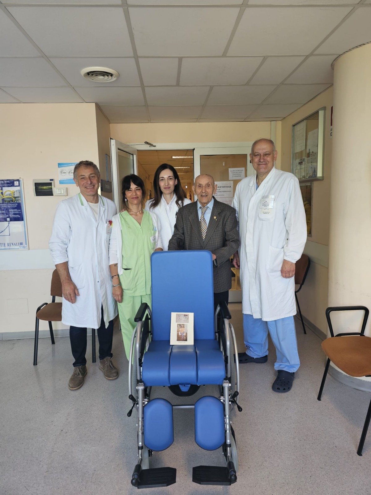 img title Un gruppo di quattro operatori sanitari in camice bianco e divisa verde posa sorridente accanto a un uomo anziano in giacca, situati nel corridoio di un reparto ospedaliero. Al centro, in primo piano, è presente una nuova poltrona medica per emodialisi di colore blu, con un cartoncino informativo appoggiato sopra.
