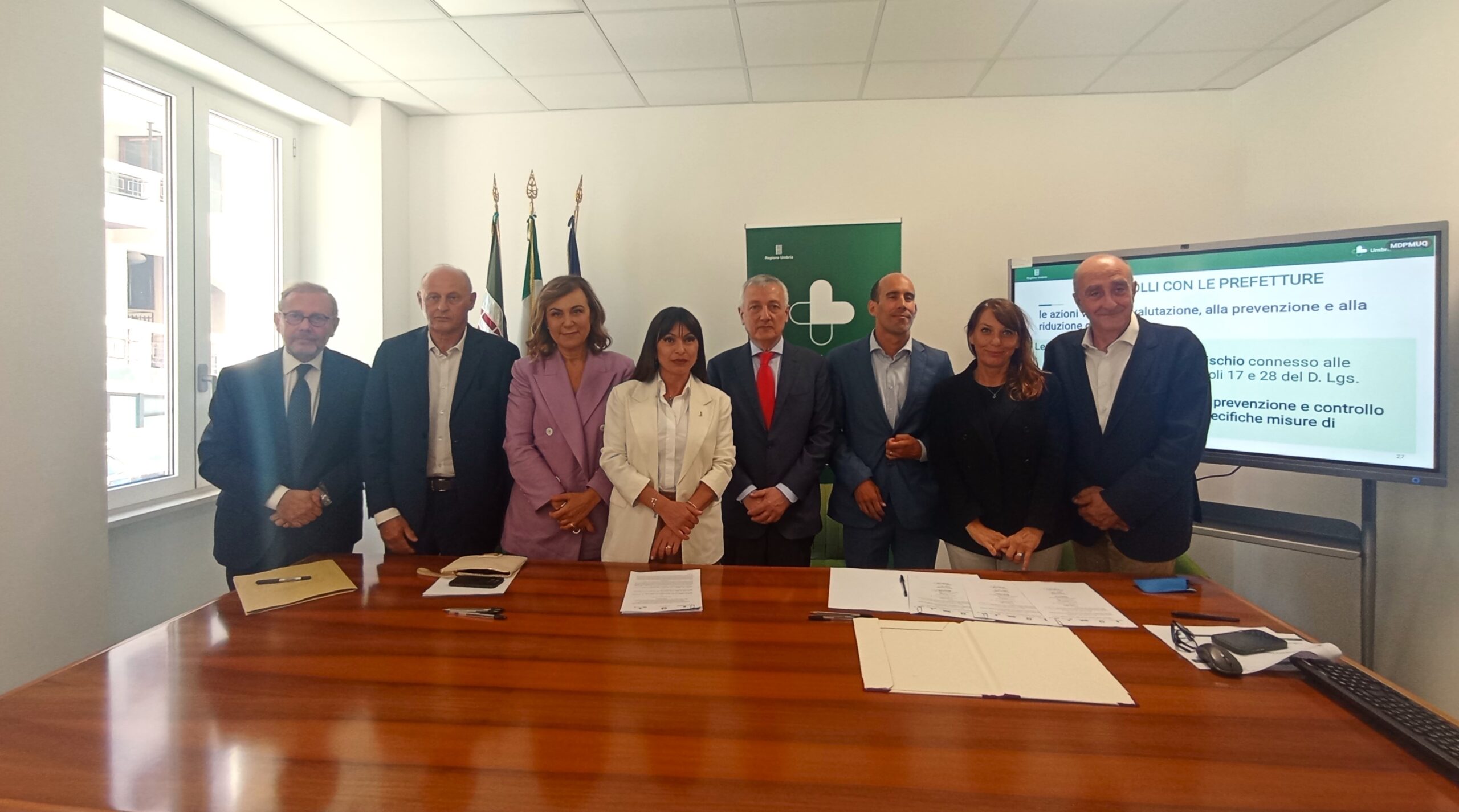 img title Fotografia istituzionale in una sala riunioni. Otto persone, sei uomini e due donne in abiti formali, posano in piedi allineate dietro a un grande tavolo in legno scuro su cui sono disposti fogli, faldoni e penne. Sullo sfondo si trovano le bandiere istituzionali, un pannello verde con un logo sanitario bianco e uno schermo digitale che mostra delle slide intitolate 'PROTOCOLLI CON LE PREFETTURE'