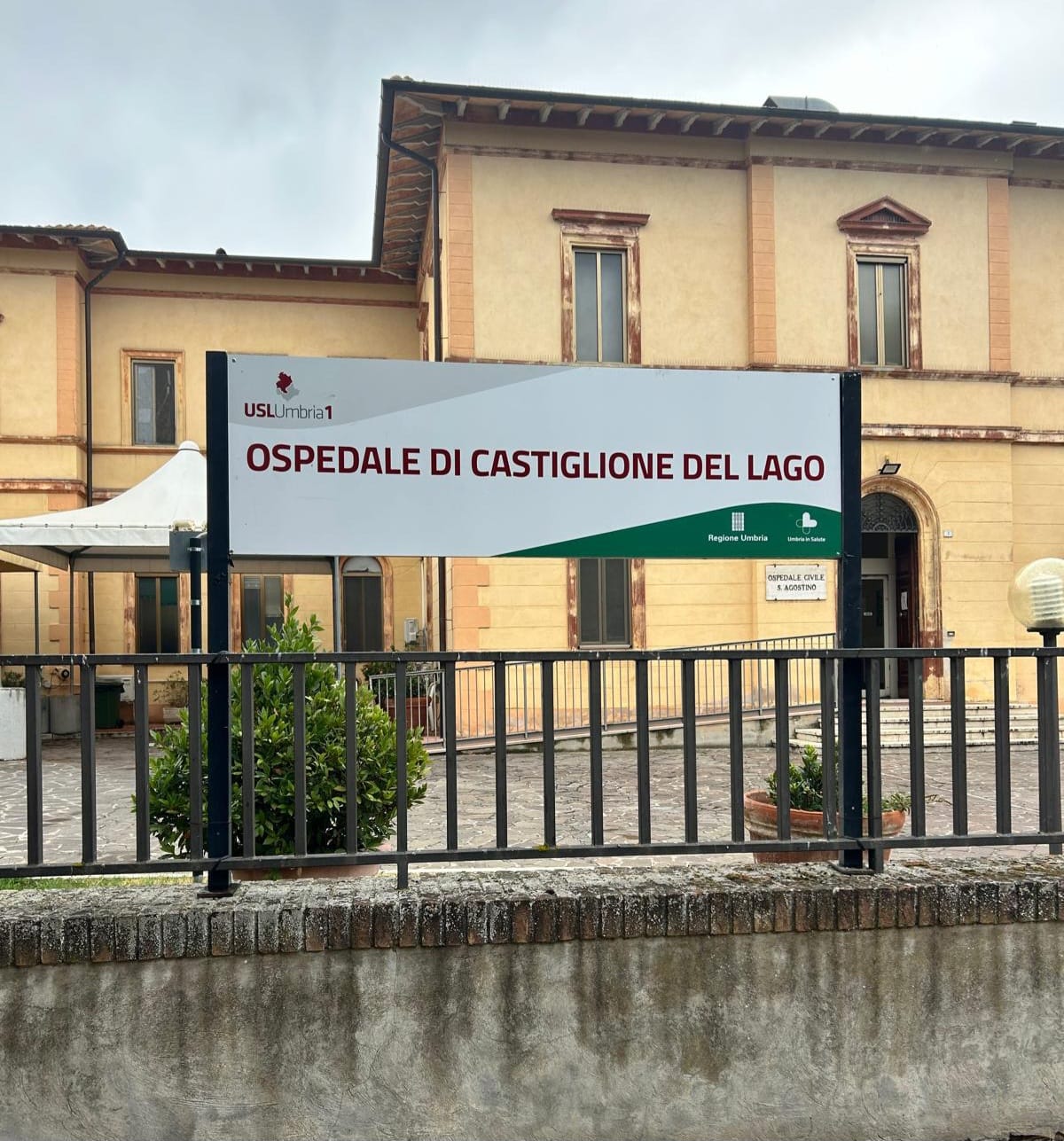 img title Cartello all'ingresso dell'Ospedale di Castiglione del Lago con loghi istituzionali e l'edificio ospedaliero sullo sfondo.