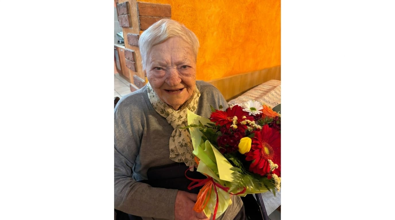 img title Primo piano di Bruna Casciari, donna di 101 anni, sorridente mentre tiene in mano un mazzo di fiori colorati con gerbere rosse e margherite. La donna indossa un maglione grigio e un foulard chiaro annodato al collo
