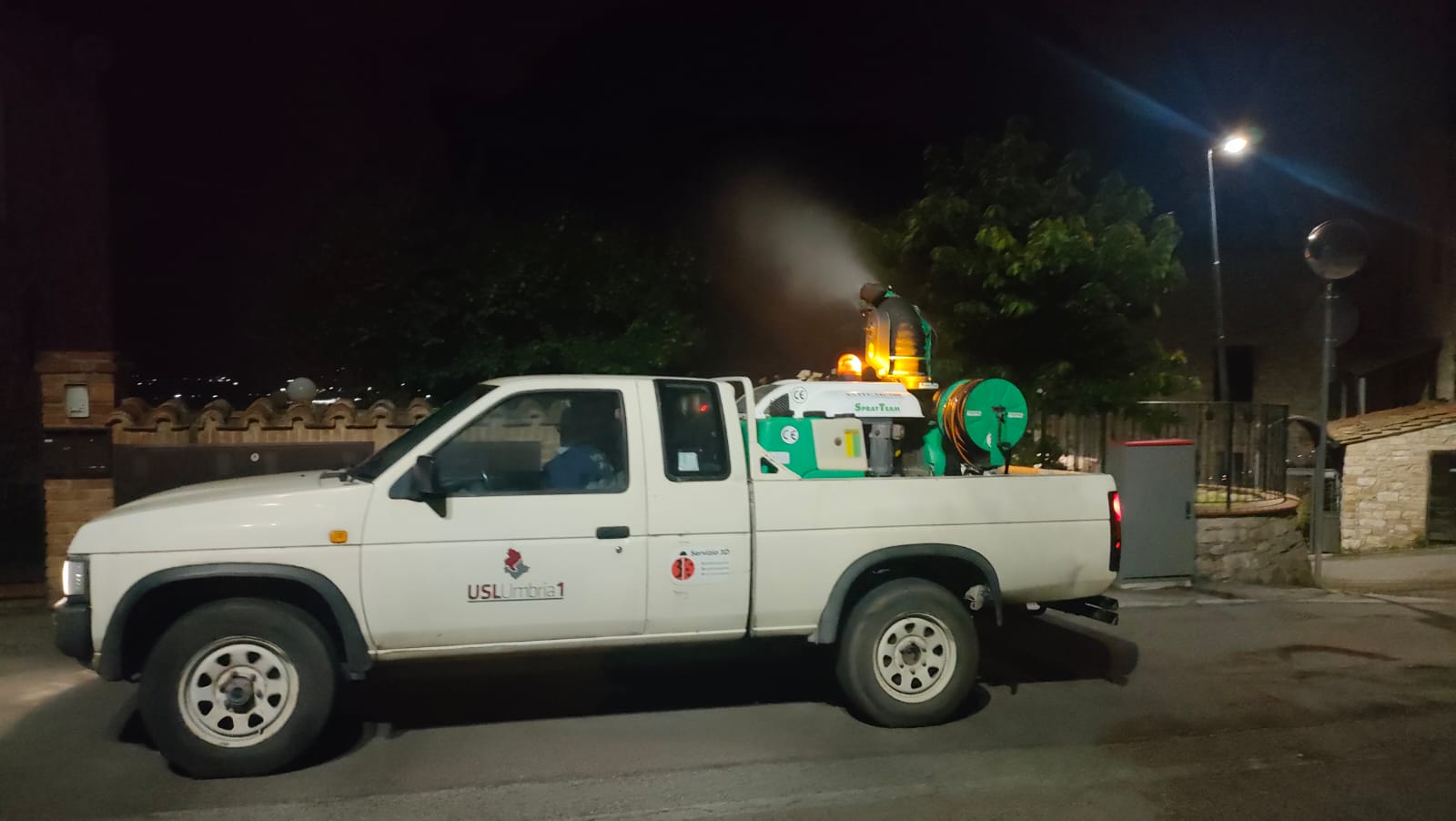 Un pick-up bianco dell'Azienda USL Umbria 1 in servizio notturno lungo una strada urbana. Sul cassone posteriore è montato un macchinario nebulizzatore in funzione che eroga una nube spray per la disinfestazione adulticida contro le zanzare.