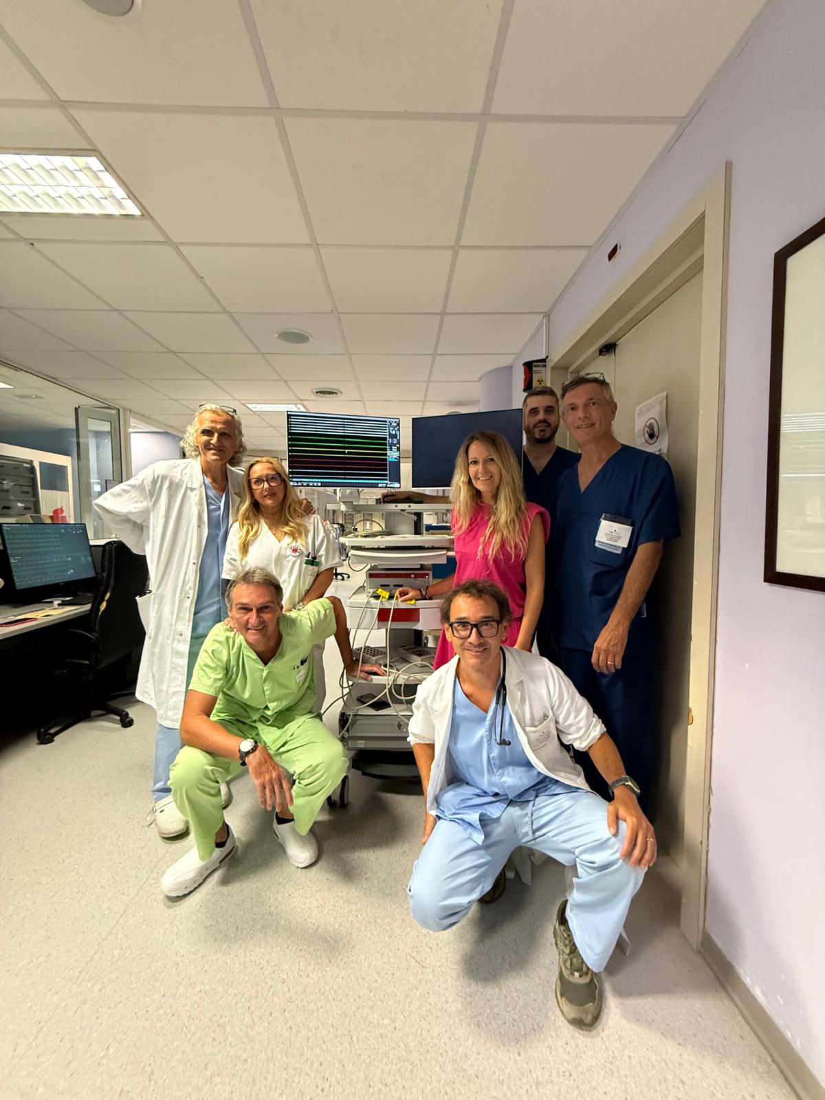 img title Équipe medica del reparto di cardiologia in posa accanto a una colonna di apparecchiature elettrocardiografiche.
