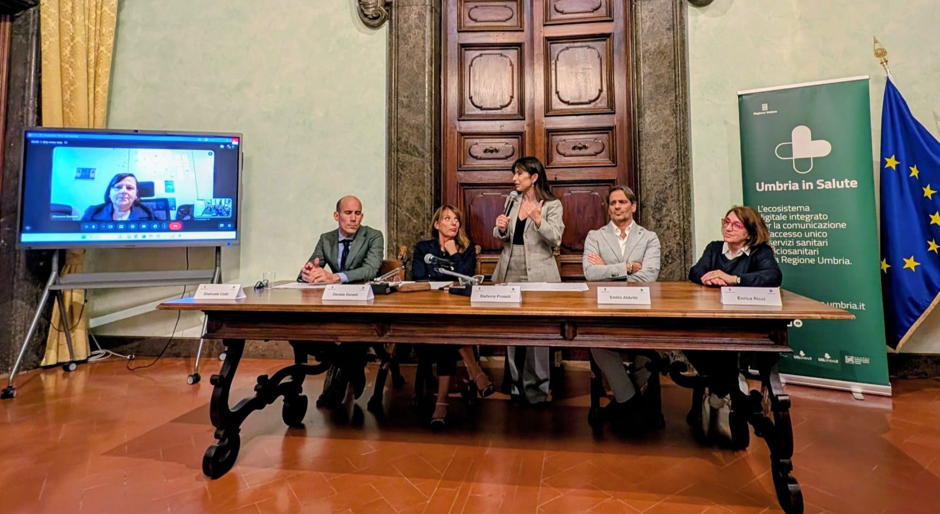 img title Tavolo dei relatori a Palazzo Donini a Perugia durante la presentazione del numero unico 116117. Al centro la presidente della Regione Umbria Stefania Proietti parla al microfono; al suo fianco da sinistra Emanuele Ciotti (DG Usl Umbria 1) e Daniela Donetti (Direttrice regionale Salute e Welfare), e sulla destra Emilio Paolo Abbritti ed Enrica Ricci. Sullo schermo a sinistra è collegata online Giovanna Giannetti del Ministero della Salute.