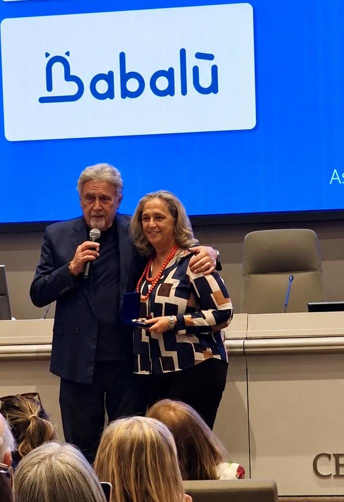 img title Premiazione istituzionale sul palco con oratore al microfono e una donna che mostra un riconoscimento davanti al logo Babalù.