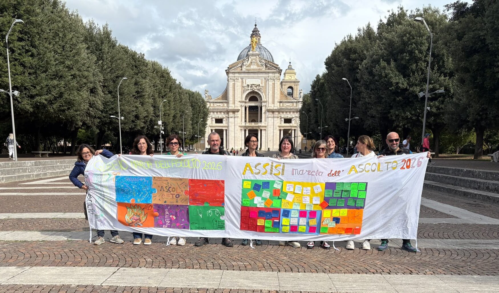 Un gruppo di partecipanti alla Marcia dell'Ascolto sostiene uno striscione bianco recante la scritta 'ASSISI marcia dell'ASCOLTO 2026', decorato con disegni e biglietti colorati. Sullo sfondo si erge la Basilica di Santa Maria degli Angeli ad Assisi.