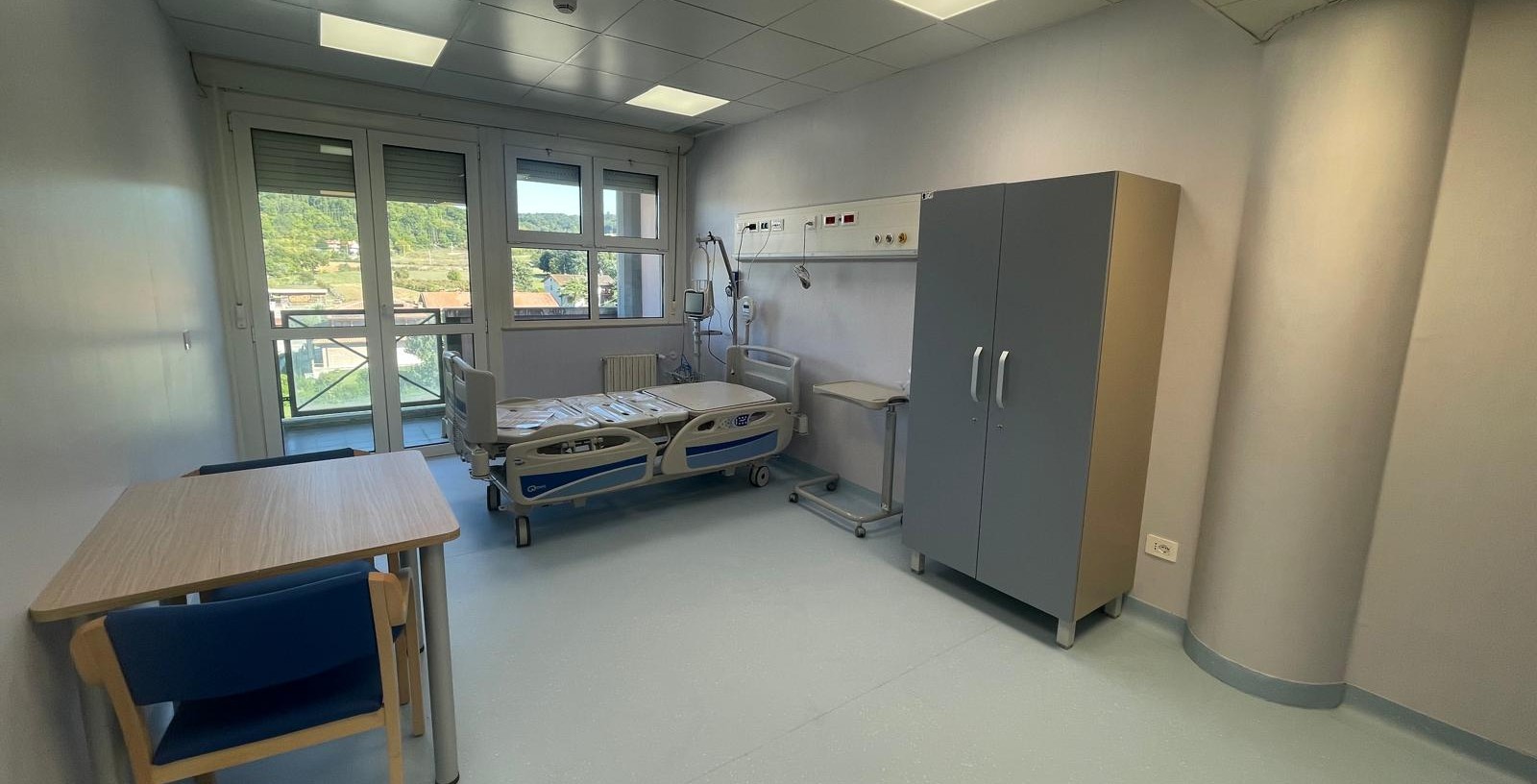 img title Interno di una stanza di degenza moderna dell'area di terapia semintensiva. Sulla destra si trova un letto di degenza elettrico articolato con testata e barre pensili mediche a parete, affiancato da un armadio grigio a due ante e un tavolino servitore. Sulla sinistra sono disposti un tavolo in legno e sedie blu. Sullo sfondo, una grande finestra e porta finestra mostrano una vista sulle colline.