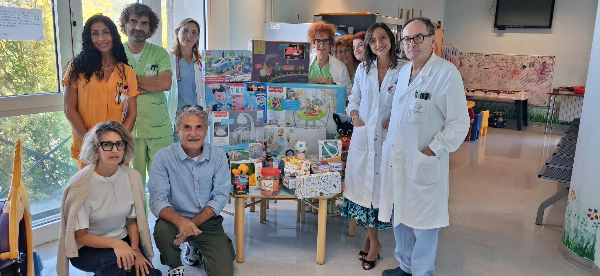 img title Foto di gruppo con il personale sanitario della Pediatria e la direzione dell'Ospedale di Città di Castello riuniti attorno a un tavolo ricolmo di giocattoli donati. Al centro sono posizionati numerosi giochi da tavolo e scatole regalo; al lato destro si trovano la direttrice f.f. del presidio Gioia Calagreti e il direttore della Pediatria Guido Pennoni in camice bianco insieme agli operatori di reparto.